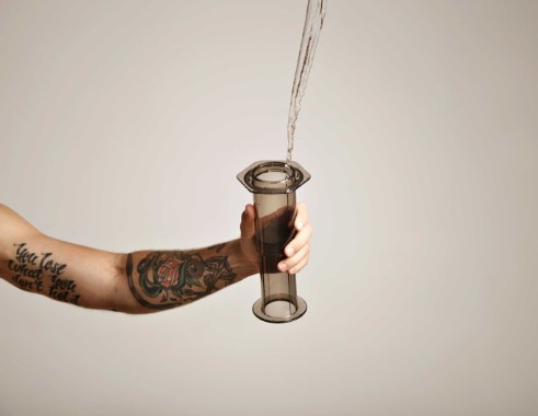 Tattooed arm holding an aeropress that water is being poured into