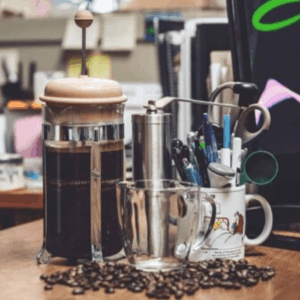 Coffee beans on desk in front of French press, cups and coffee grinder