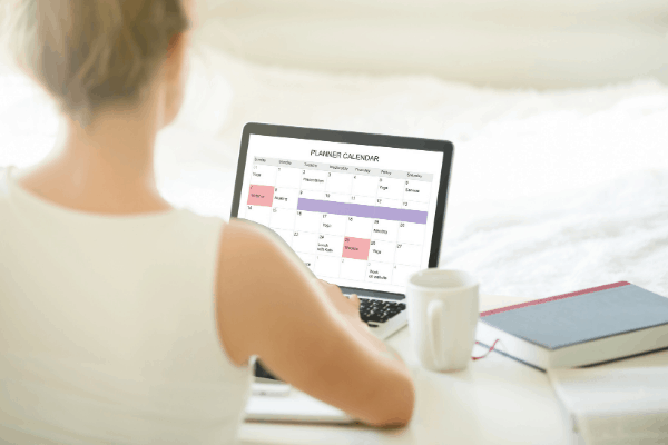 back of a woman in white working on her laptop calendar, cup of coffee and a book