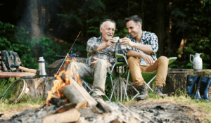 Two men in lawnchairs behind a campfire, clinking coffee cups