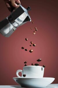 Bialetti Moka pot pouring coffee beans into a white cup and saucer, in front of a red background