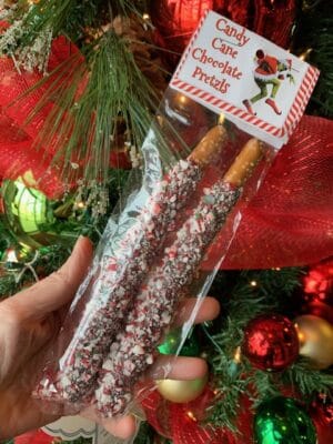 A bag of two chocolate dipped, candy cane covered pretzels in front of a christmas tree