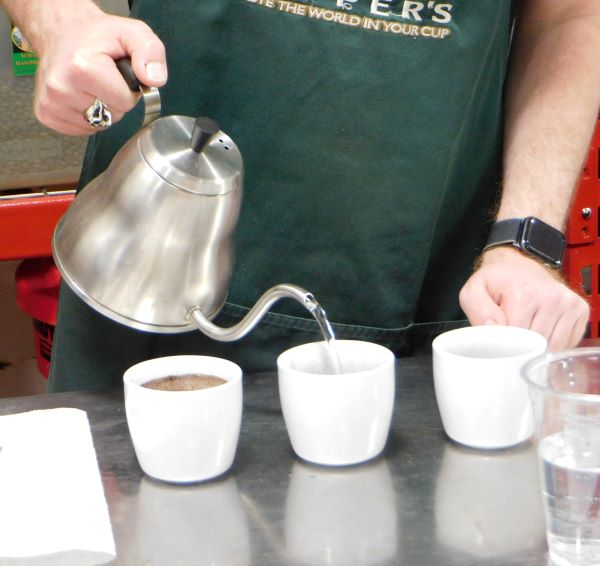 a hand holding a goose necked kettle pouring hot water over coffee grounds in a white cup as part of the coffee cupping process