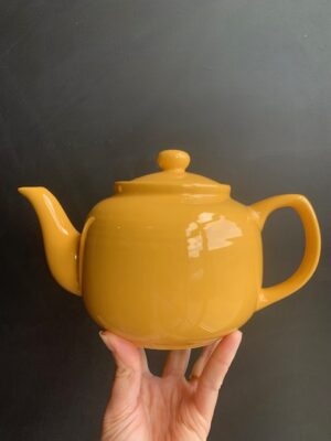 Yellow 6 cup Windsor Teapot being held up by a hand in front of a blackboard