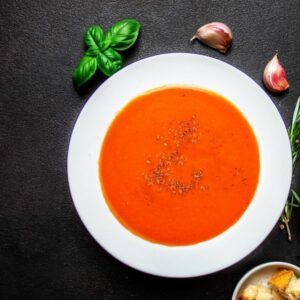 A bowl of creamy tomato soup with basil leaves and garlic cloves