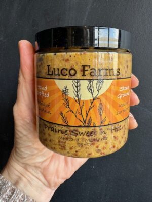 Luco Farms Prairie Sweet n' Hot mustard in a plastic jar, being held by a hand