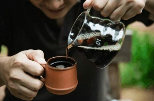 Man pouring coffee from a small carafe to a brown mug.