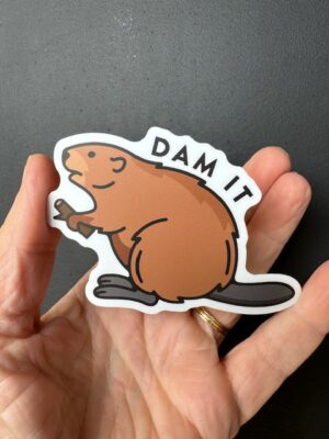 Hand holding a sticker with a beaver holding a stick and the words "Dam it"