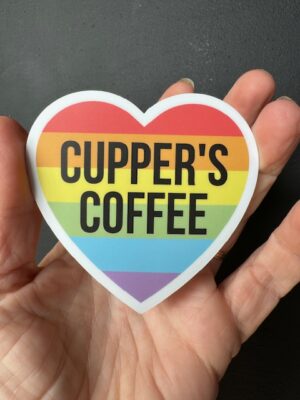 Hand holding a sticker with a rainbow heart with words 'Cupper's Coffee'