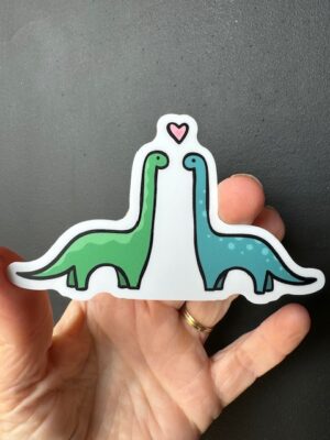 Hand holding a sticker with a green brontosaurus dinosaur and blue brontosaurus dinosaur facing each other with a pink heart above their heads