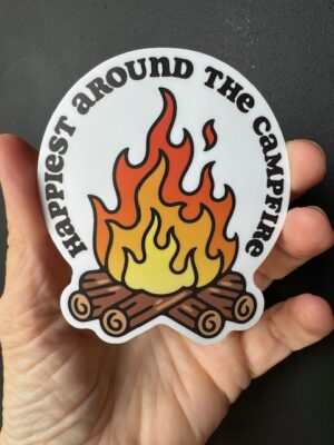 Hand holding a sticker with fire on logs and words 'Happiest Around the Campfire'