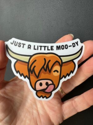 Hand holding a sticker with a head of a Highland cow licking its nose and the words "Just a little Moo-dy"