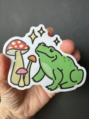 Hand holding a sticker with three clustered mushrooms, gleams and a green frog