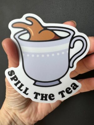 Hand holding a sticker with a tea cup slopping tea, and the words "Spill the tea" under it