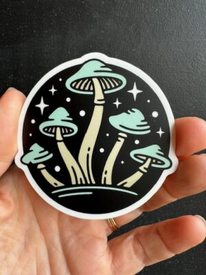 Hand holding a sticker with blue capped mushrooms on a starry night sky background