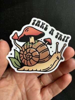 Hand holding a sticker with a snail with mushrooms on it, and text "Take a trip"