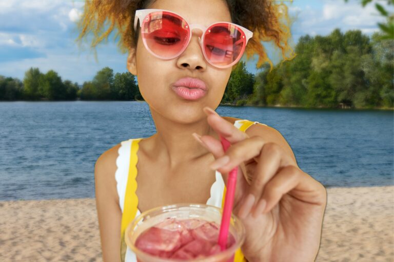 A woman with sunglasses and swimsuit at a beach holding and looking at a cold drink with a straw
