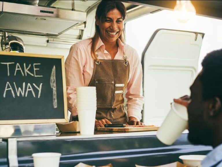 a smiling woman in an leather apron in a food truck taking an order from a man holding a paper coffee cup.