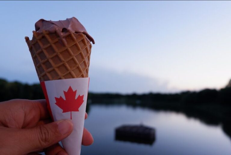 A hand holding an ice cream cone with a Canada wrapper on the bottom, all in front of a blurry lake background in early evening