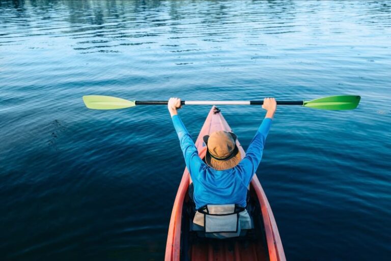 A kayaker in a red kayak, dressed in blue holding up a green bladed paddle on calm water