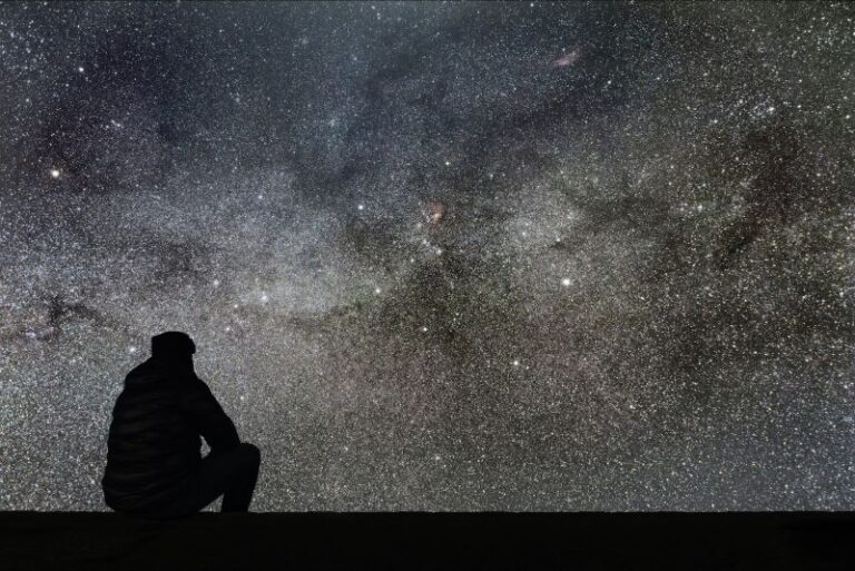 a man in silhouette sitting looking at a vast backdrop of stars in the night sky