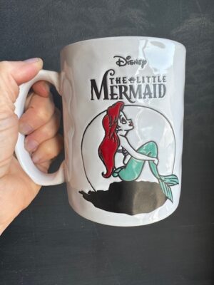 Little Mermaid Mug in white with classic image of Ariel holding her tail and looking up. Wax removed style of glaze over graphic.