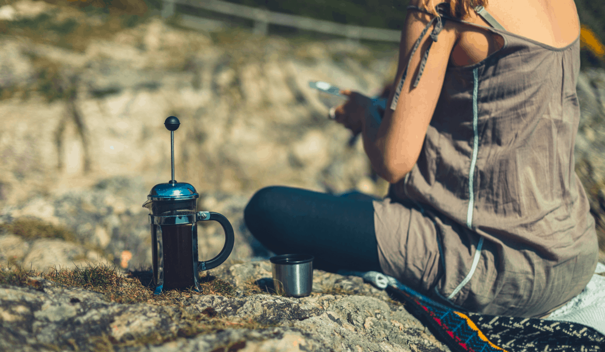 Woman sitting on a woven blanket on a large rock outcropping, with a French coffee press beside her.