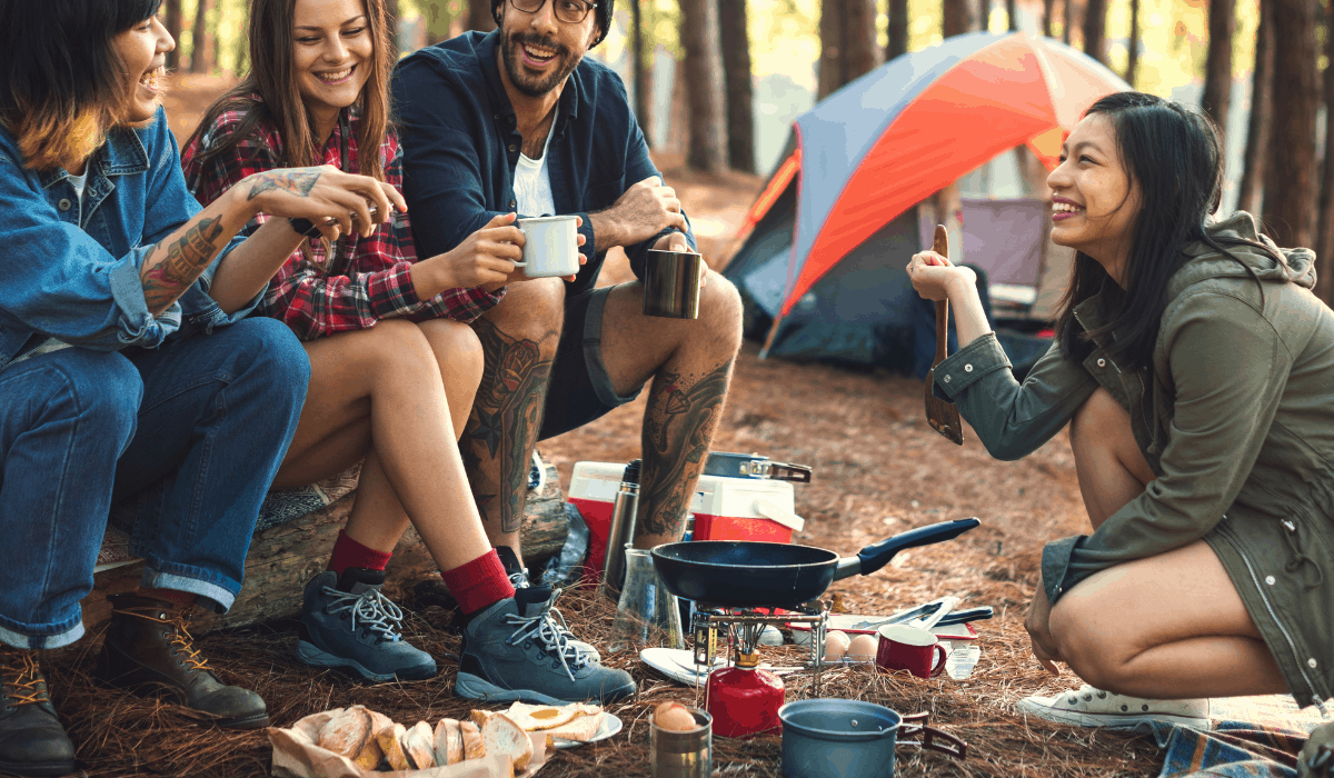 In a forest, three people sitting and one crouching around a backpacking cookstove with camping gear at their feet and a tent in the background