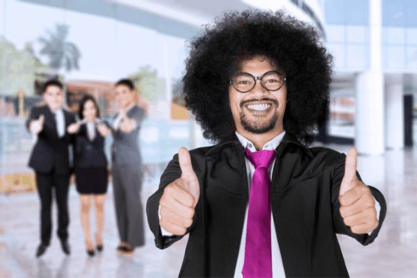 Curly-haired smiling man with bright pink tie and black jacket with two thumbs up, three blurred people behind